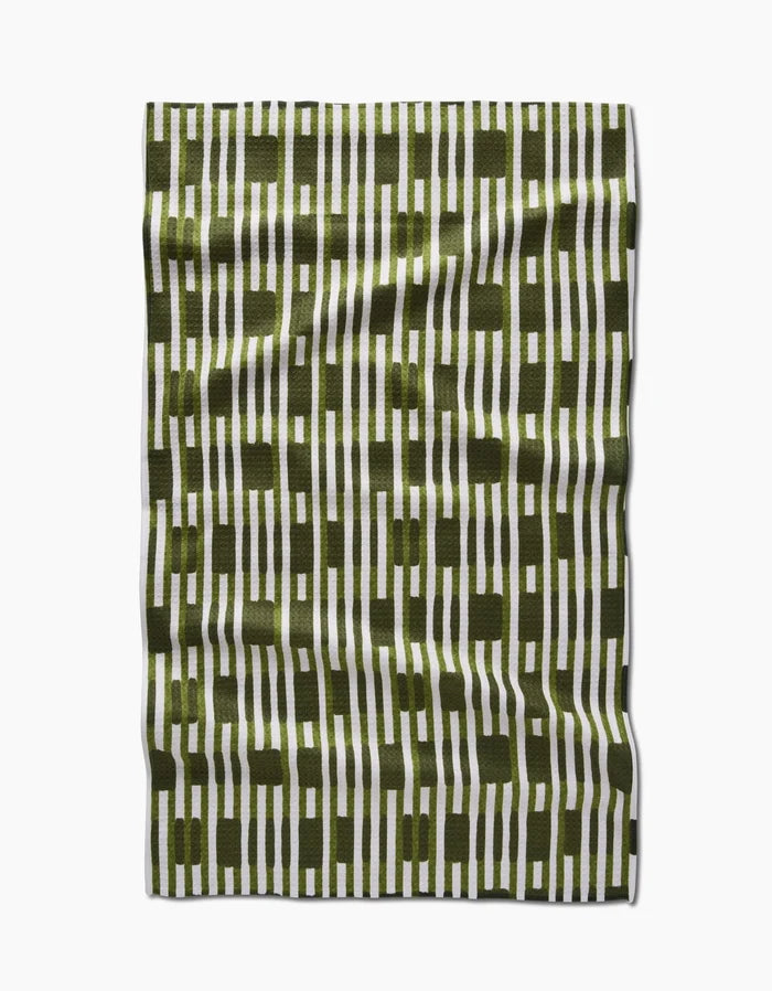 The GEOMETRY Clover Stripe tea towel features vertical olive green and white stripes with intersecting olive green blocks, shown laid flat on a white background with a slightly wrinkled fabric texture.