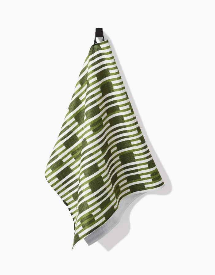 The Clover Stripe kitchen towel by GEOMETRY, featuring a green and white geometric pattern, hangs from a black hook against a white background.