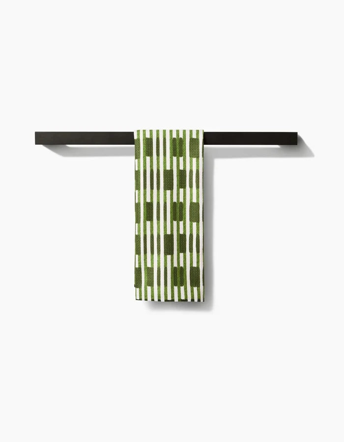 The GEOMETRY Clover Stripe recycled kitchen towel, featuring a green and white pattern, hangs neatly over a sleek black horizontal towel bar against a plain white background.