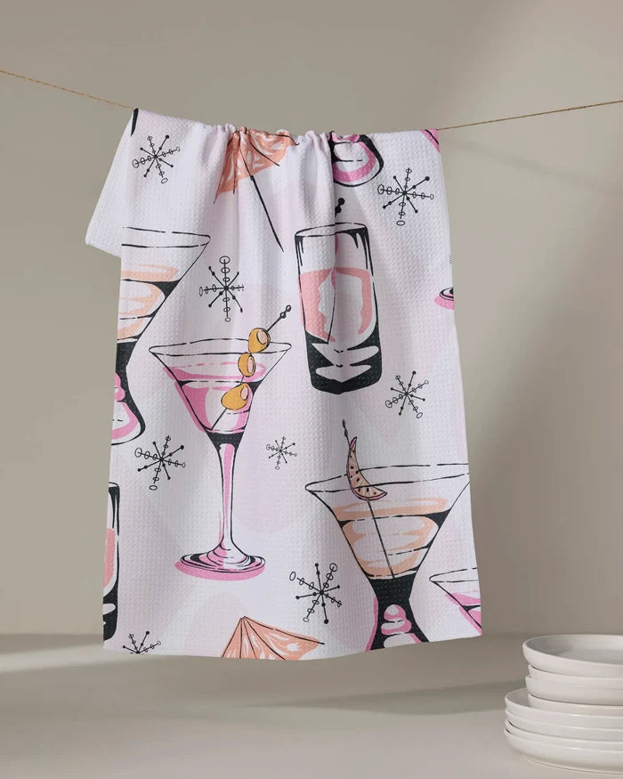 The "Cocktail Hour" tea towel by Julianne Haness, with a retro cocktail print of martini and highball glasses, olives, and swizzle sticks, hangs above stacked white plates.