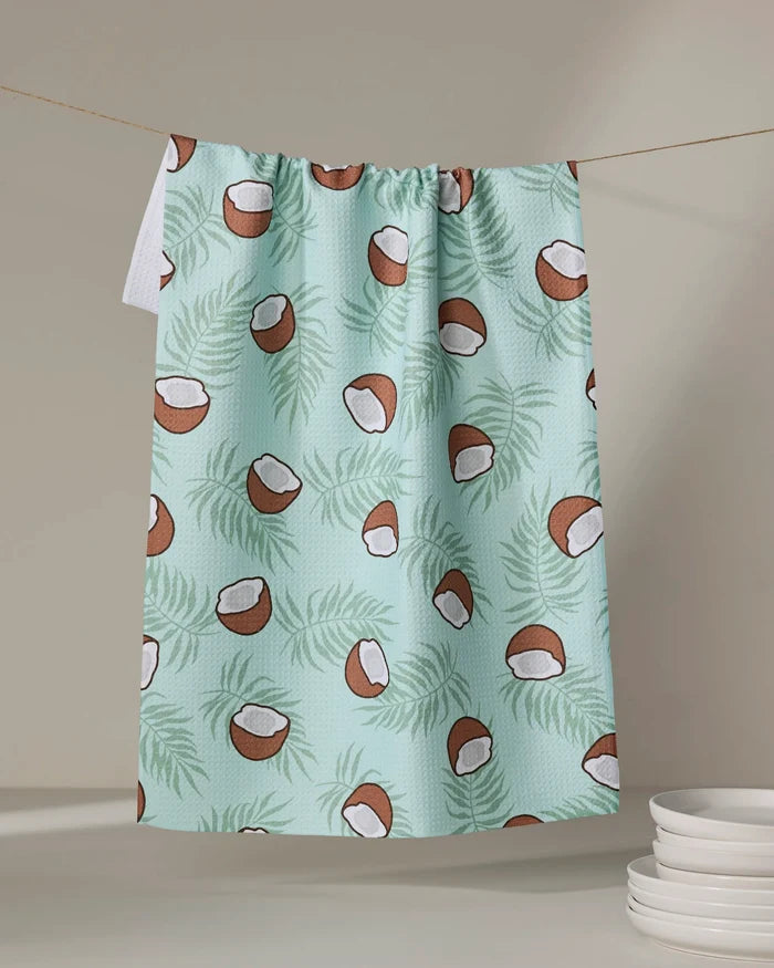 The Purely Kaylie Coconut Waves recycled kitchen towel, featuring coconut halves and green palm leaves on light blue, hangs on a line. A stack of white plates appears in the lower right corner against a neutral background.