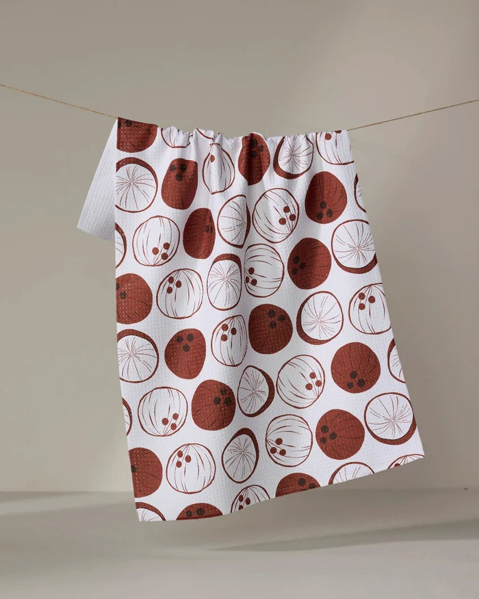 The "Coconutz About You" tea towel by Sara Mari features red and brown circular fruit slice patterns on white, crafted from ultra-absorbent fabric, and hangs on a clothesline against a plain, neutral background.
