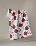 The "Coconutz About You" tea towel by Sara Mari features red and brown circular fruit slice patterns on white, crafted from ultra-absorbent fabric, and hangs on a clothesline against a plain, neutral background.