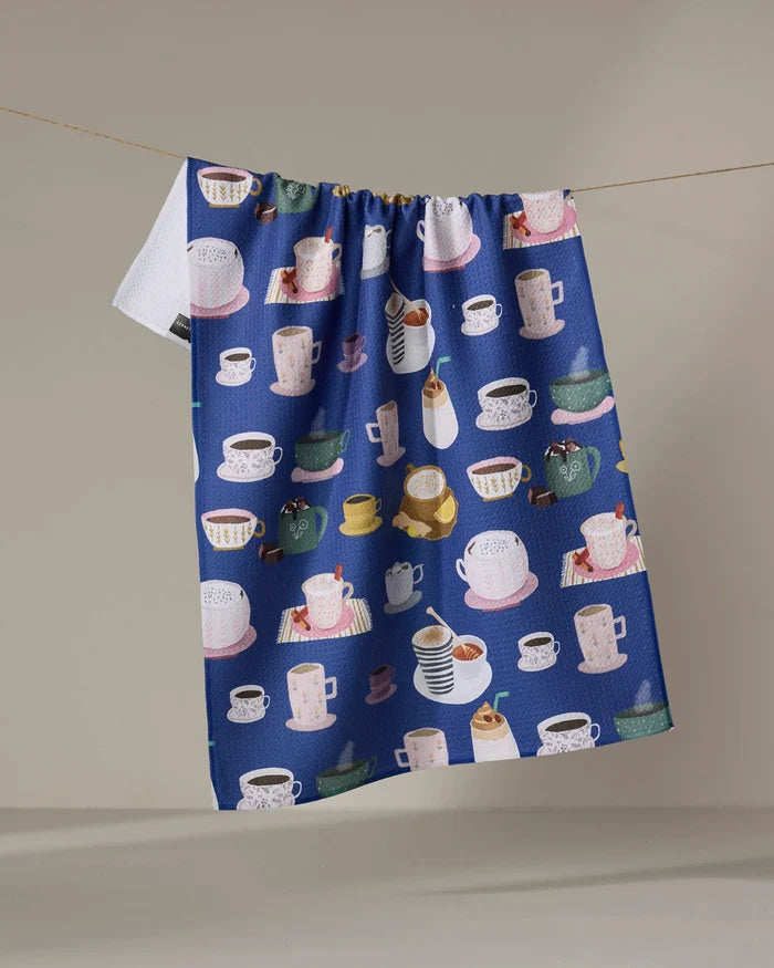 The Coffee Drinks kitchen towel by Cottonflower Studio is blue, made from recycled materials, and features colorful mugs, cups, teapots, and desserts hanging on a clothesline against a neutral background.