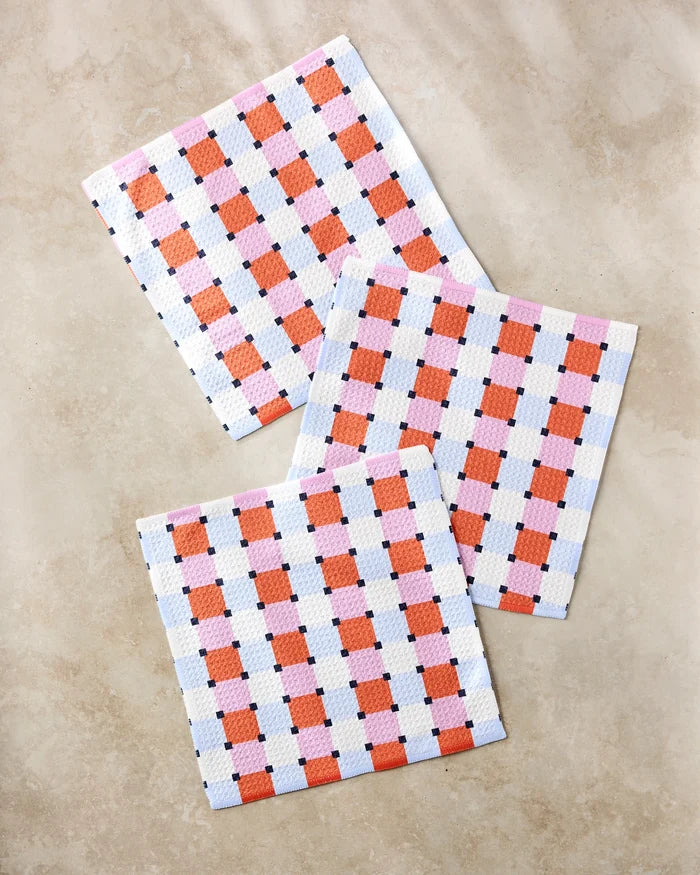 Three Color Block Grid cloths by Claire Iglesias, featuring red, blue, and white checks, are displayed on a beige textured surface; this eco-friendly dishcloth set is crafted from recycled materials for a sustainable kitchen upgrade.