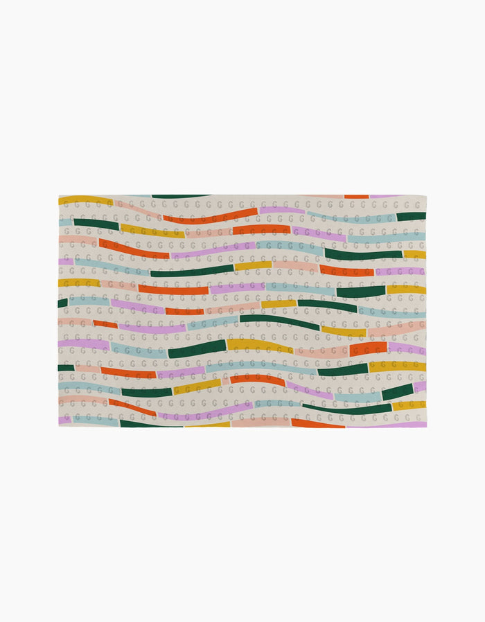 The Geometry Colorful Currents rug is rectangular and eco-friendly, made from post-consumer recycled materials. It features wavy green, yellow, orange, pink, and purple stripes with repeating “G” patterns on a beige background.