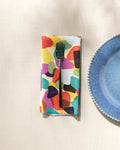 A silver fork sits on GEOMETRY’s Colorful Pebbles recycled napkin, placed beside a blue scalloped-edge plate on a light textured surface.