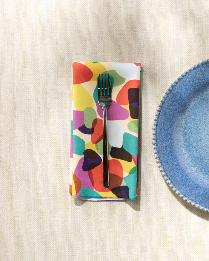 A silver fork sits on GEOMETRY’s Colorful Pebbles recycled napkin, placed beside a blue scalloped-edge plate on a light textured surface.