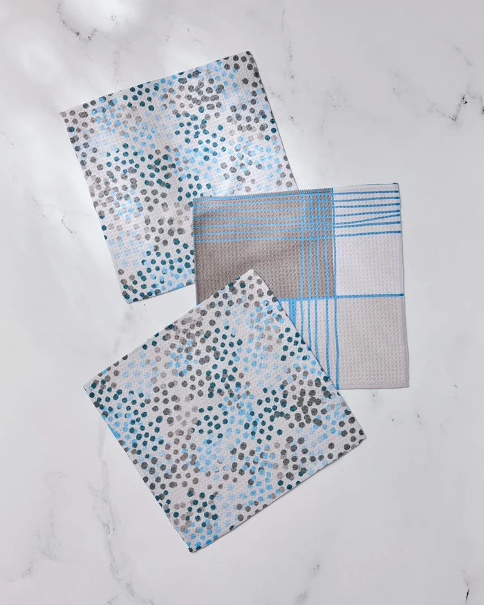 Three Geometry dishcloths from the Cool Blocks and Dots set are displayed on a white marble surface—two featuring blue, grey, and black dot patterns, and one with a blue plaid design—all made from recycled materials.