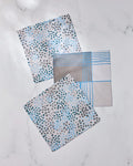 Three Geometry dishcloths from the Cool Blocks and Dots set are displayed on a white marble surface—two featuring blue, grey, and black dot patterns, and one with a blue plaid design—all made from recycled materials.