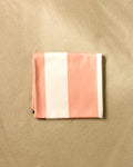 The GEOMETRY Coral Block Stripes beach blanket, featuring wide pink and white stripes and crafted from quick-drying recycled materials, rests on smooth light sand.