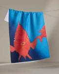 The Trey Jolley "Coral Glow" is a blue, quick-drying tea towel made from recycled materials, featuring a surprised red fish illustration, shown hanging on a clothesline against a neutral background.