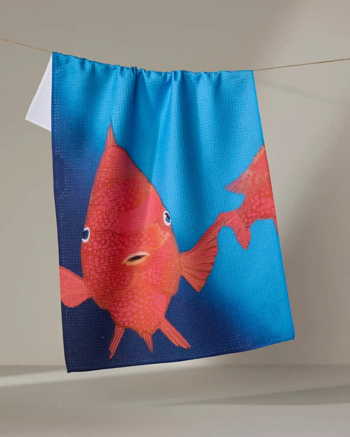 The Trey Jolley "Coral Glow" is a blue, quick-drying tea towel made from recycled materials, featuring a surprised red fish illustration, shown hanging on a clothesline against a neutral background.