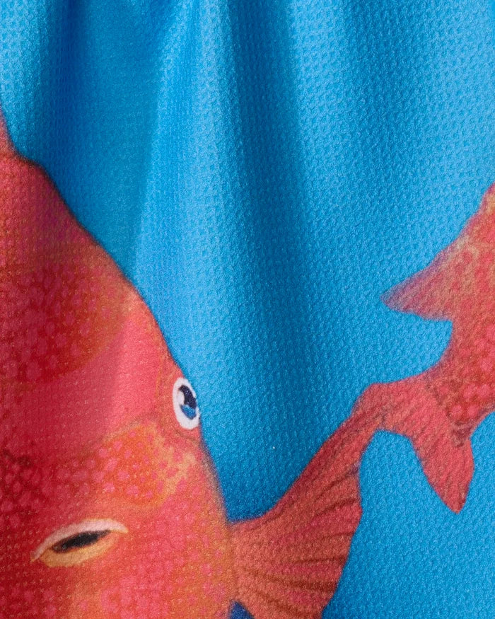 A close-up of the Trey Jolley "Coral Glow" tea towel, made from recycled materials, shows a red-orange fish with a large eye and detailed fins on a textured blue background. The fabric appears slightly wrinkled.