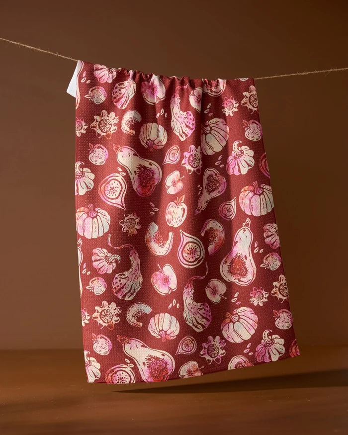 The Geometry Cosecha tea towel, ultra-absorbent and maroon with a pink and white fig and fruit pattern, hangs on a clothesline against a brown background.