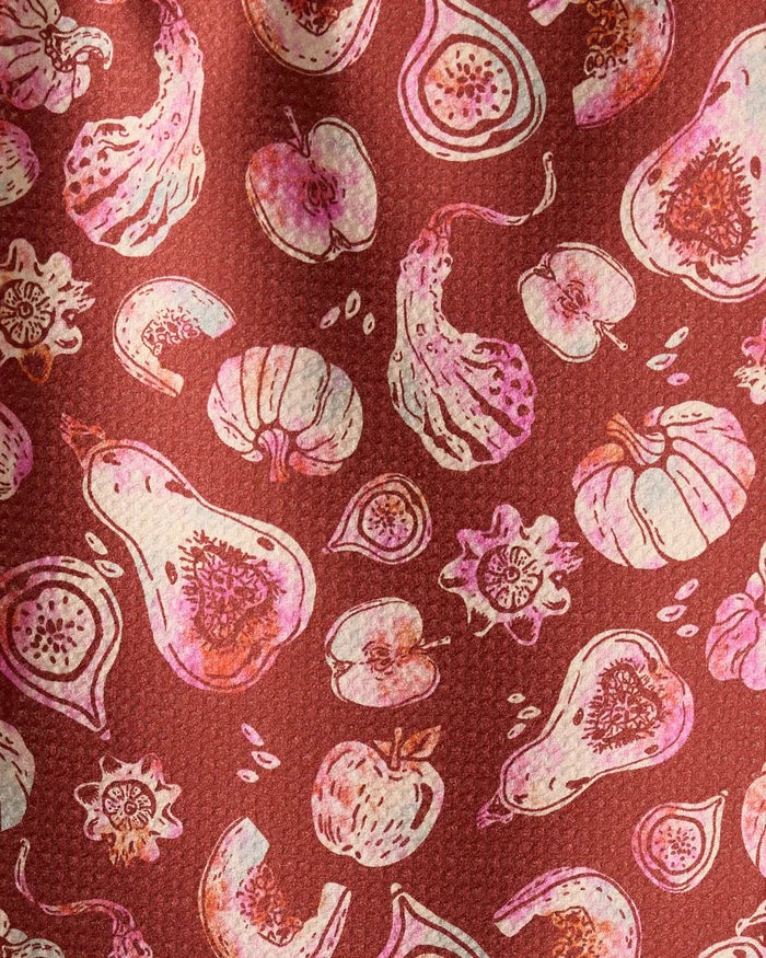 Geometry’s Cosecha bar towel features illustrations of fruits and vegetables—like gourds, figs, pumpkins, and apples—in pink and cream on a textured reddish-brown background. Made from ultra-absorbent recycled materials.