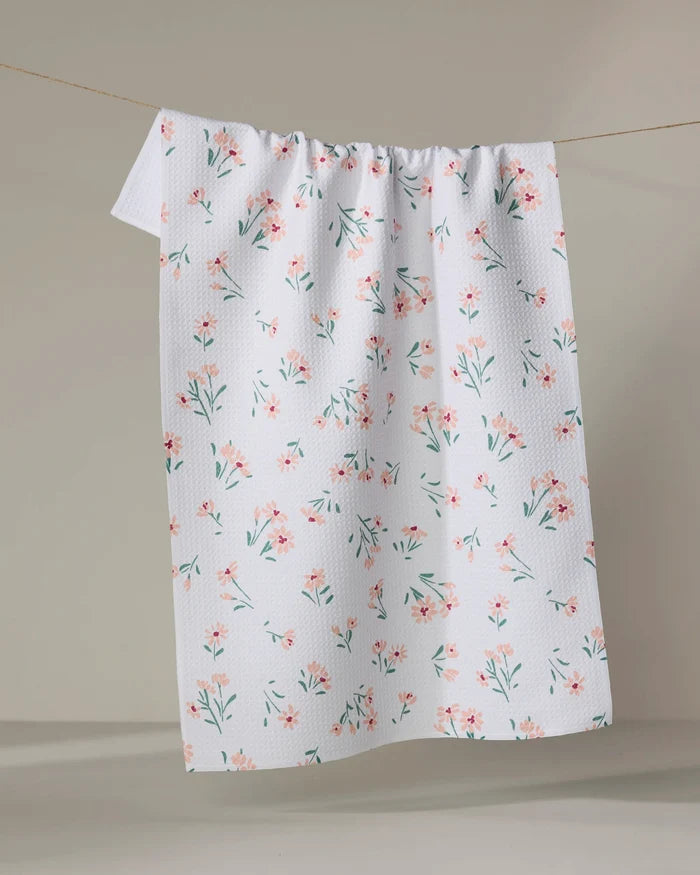 The Cottage Garden tea towel by Hufton Studio, made from recycled materials and featuring a white textured fabric with small pink and green flowers, hangs on a clothesline against a plain beige background.