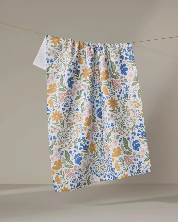 The Danelys Sidron Cottage Meadow Pastel kitchen tea towel, featuring a blue, yellow, pink, and green floral pattern, hangs on a clothesline against a plain beige background.
