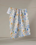 The Danelys Sidron Cottage Meadow Pastel kitchen tea towel, featuring a blue, yellow, pink, and green floral pattern, hangs on a clothesline against a plain beige background.