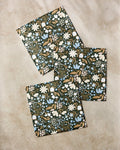 Three "Cottage Meadow Sepia" rectangular notebooks by Danelys Sidron, with dark covers featuring white, blue, and tan floral designs, are arranged on a beige surface, echoing the style of eco-friendly dishtowels.
