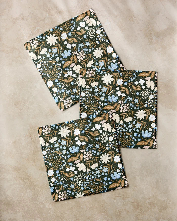 Three "Cottage Meadow Sepia" rectangular notebooks by Danelys Sidron, with dark covers featuring white, blue, and tan floral designs, are arranged on a beige surface, echoing the style of eco-friendly dishtowels.