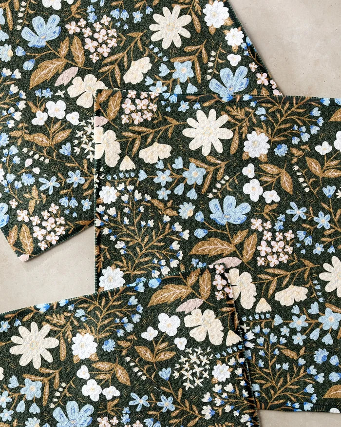 Cottage Meadow Sepia by Danelys Sidron: recycled paper sheets with a blue, white, and gold floral pattern on dark green. Overlapping sheets show scattered flowers and leaves arranged on a light surface.