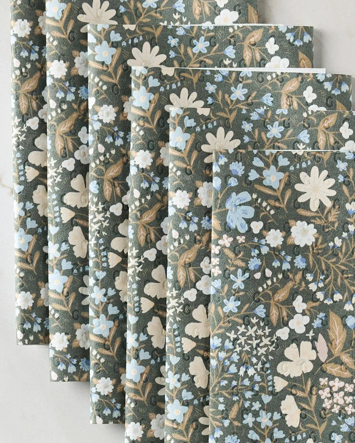 Danelys Sidron’s Cottage Meadow Sepia features several sheets of decorative, recycled paper towel alternatives with a dense blue, white, and tan floral pattern on a dark green background, fanned out on a light surface.