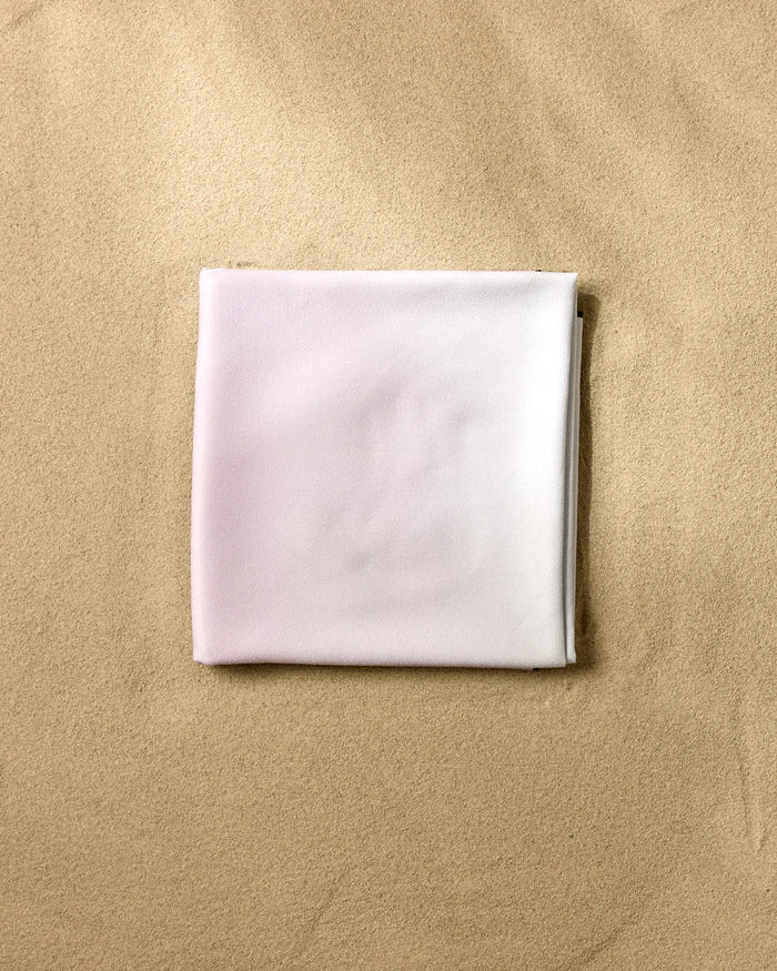 A neatly folded GEOMETRY Cotton Candy beach blanket, made from recycled materials, rests on smooth, light brown sand.