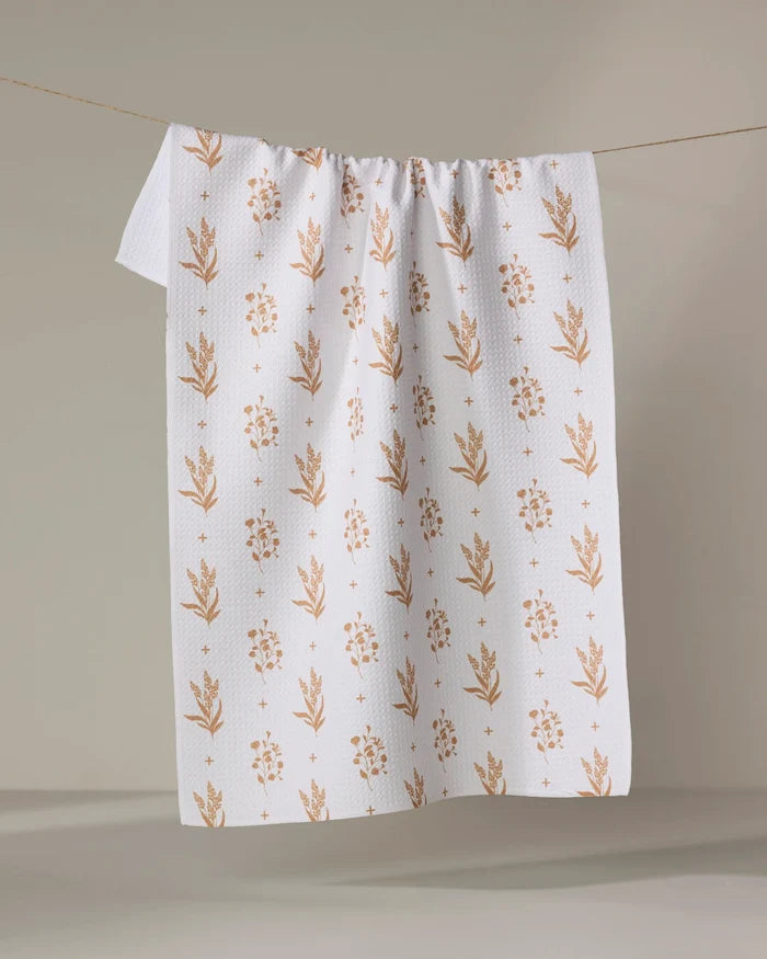 The "Country Road" kitchen towel by Charlie Rowan Designs features a textured white surface with light brown floral patterns, made from recycled materials, and hangs on a string against a neutral background.
