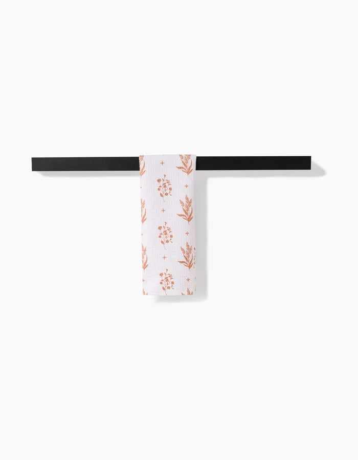 The Country Road bar towel by Charlie Rowan Designs features an orange floral pattern on white Geoweave fabric and hangs from a sleek black bar—a modern, versatile kitchen accessory that complements any contemporary space.
