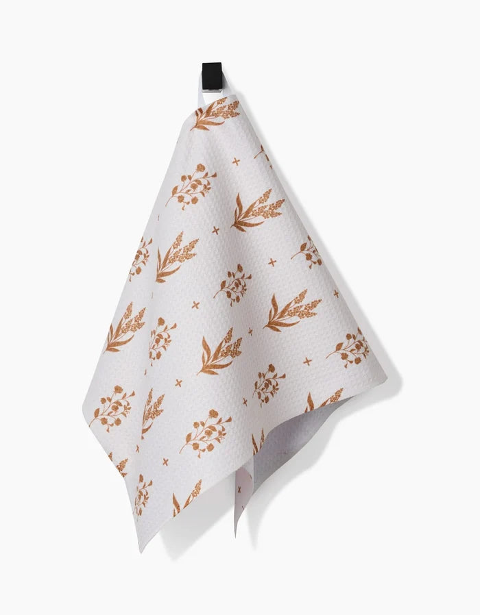 The Country Road bar towel by Charlie Rowan Designs features a brown floral and leaf pattern on white Geoweave fabric, hanging from a black hook against a white backdrop—a stylish and functional kitchen accent.
