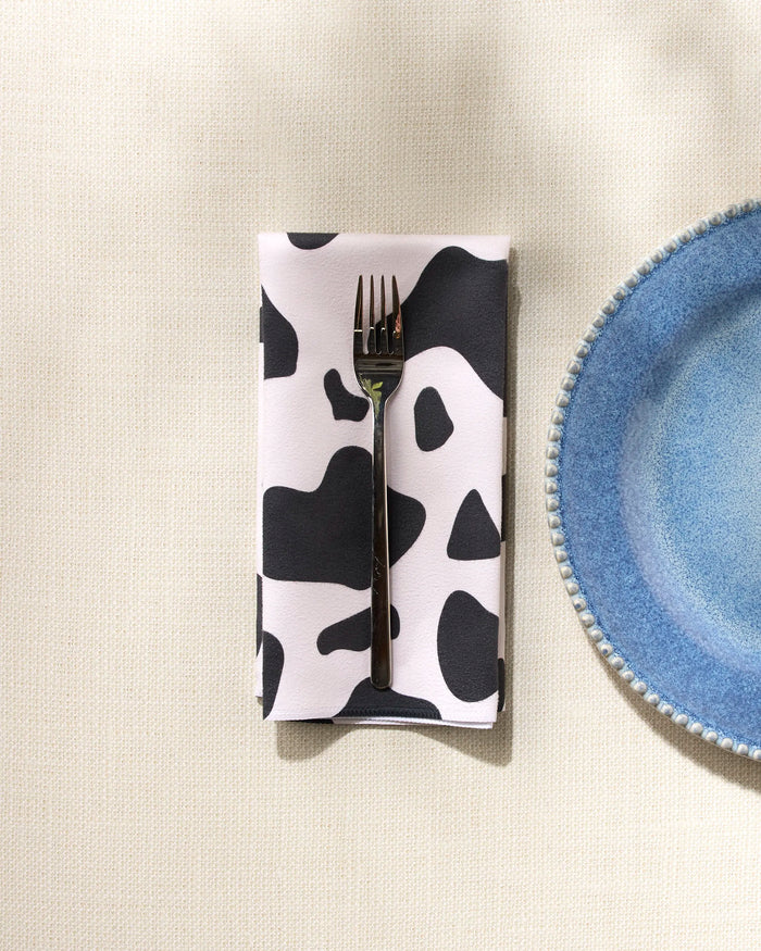 A metal fork rests on a GEOMETRY Cow Print Black eco-friendly napkin beside a blue speckled ceramic plate with a textured rim, all set atop a light, textured tablecloth.
