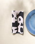 A metal fork rests on a GEOMETRY Cow Print Black eco-friendly napkin beside a blue speckled ceramic plate with a textured rim, all set atop a light, textured tablecloth.