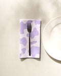 A silver fork rests on the GEOMETRY Cow Print Lilac recycled dinner napkin, featuring a lavender and white pattern, atop a light textured tablecloth beside a white plate’s edge.