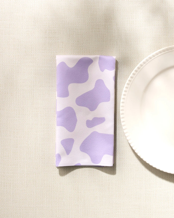 The GEOMETRY Cow Print Lilac Dinner Napkin Set, made from soft Geosuede fabric with a light purple and white cow print, is placed beside a white plate on a textured, light-colored tablecloth.