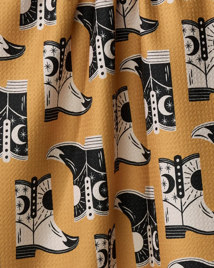 A close-up of Megan Roy’s "Cowboy Boots" quick-drying yellow fabric, featuring a repeating black and white cowboy boot pattern with moon and star motifs. The material is slightly wrinkled.