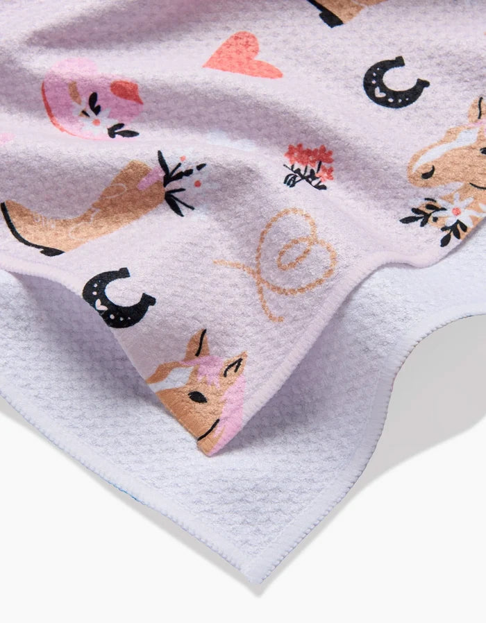 A close-up of Amberleigh Adoff’s Cowgirl Love ultra-absorbent white tea towel, featuring playful pink, brown, and black prints of cowboy boots, horses, horseshoes, lassos, hearts, and hats.