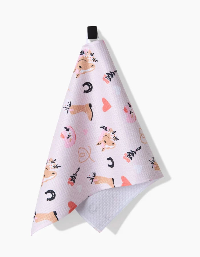Amberleigh Adoffs Cowgirl Love pink ultra-absorbent bar towel features cowgirl-themed art—cow faces, horseshoes, lassos, boots with flowers, and hearts. Hang it by the black loop to add a Western touch to your kitchen.