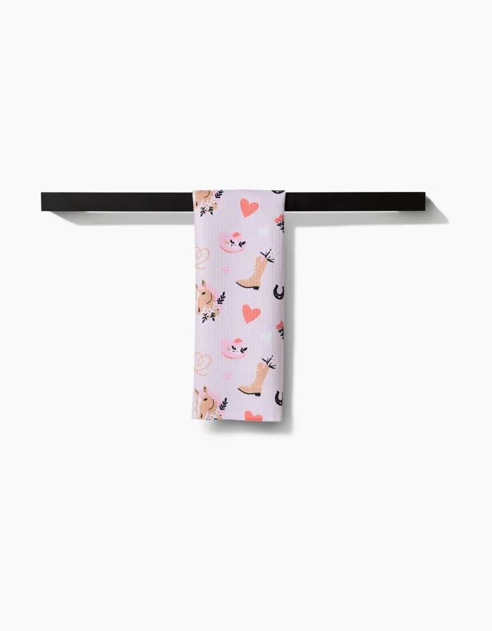 The Cowgirl Love towel by Amberleigh Adoff, featuring a pink cat, heart, and leaf print on ultra-absorbent white fabric, is neatly draped over a sleek black towel bar against a white background.
