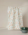 A GEOMETRY Crayon Floral recycled white kitchen towel with an orange and green floral pattern hangs on a line. Two green ceramic bowls are stacked in the bottom right corner against a beige background.