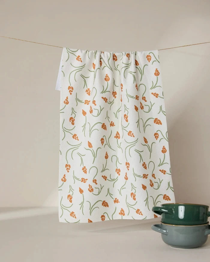 A GEOMETRY Crayon Floral recycled white kitchen towel with an orange and green floral pattern hangs on a line. Two green ceramic bowls are stacked in the bottom right corner against a beige background.