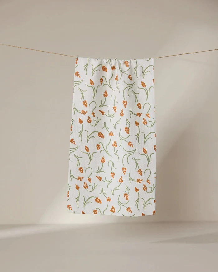 The GEOMETRY Crayon Floral ultra-absorbent white towel, featuring an orange and green floral design, hangs against a neutral beige background.