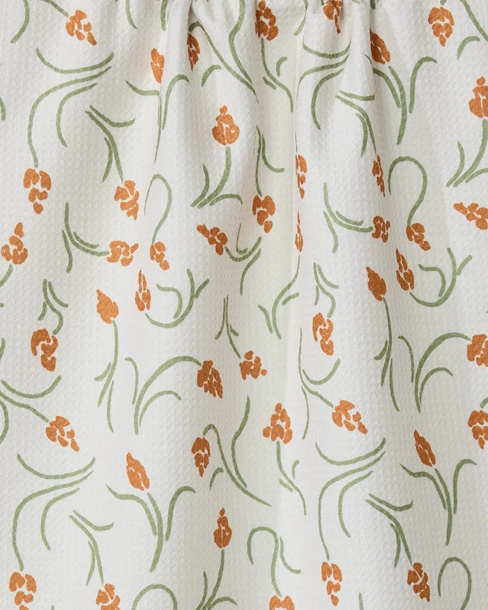 The GEOMETRY Crayon Floral tea towel features white textured fabric with orange floral clusters and green curving stems and leaves. Slightly gathered at the top, it offers style, function, and a light, airy look.