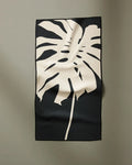 The PrintedPlant Cream Monstera is a rectangular black fitness towel made from recycled materials, featuring a large beige monstera leaf design and ultra-absorbent fabric with gentle folds, shown hanging on a plain light gray wall.