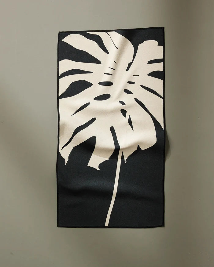 The PrintedPlant Cream Monstera is a rectangular black fitness towel made from recycled materials, featuring a large beige monstera leaf design and ultra-absorbent fabric with gentle folds, shown hanging on a plain light gray wall.