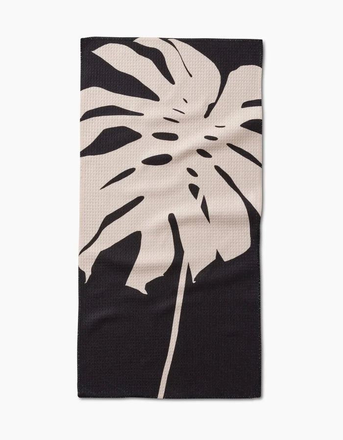 The Cream Monstera Bar Towel by PrintedPlant features a large beige monstera leaf on dark Geoweave fabric, offering superior absorbency and style.