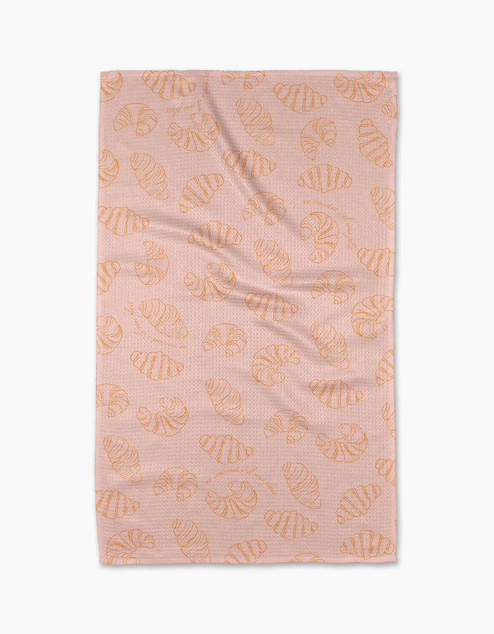 The Croissant Crush tea towel by Geometry, made from recycled materials, features orange croissants on pink and is shown laid flat against a white background.