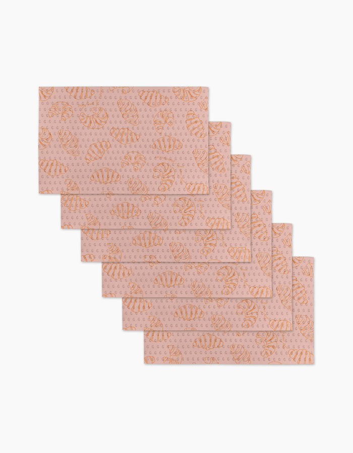 Six reusable Croissant Crush placemats by Geometry, featuring a pink background with orange croissants and polka dots, are stacked in a staggered overlap on a white surface.