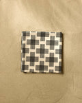 A folded Rosa Kichline Cross Hatch Micro-loop Beach Blanket in beige with a black geometric pattern rests on smooth sand.