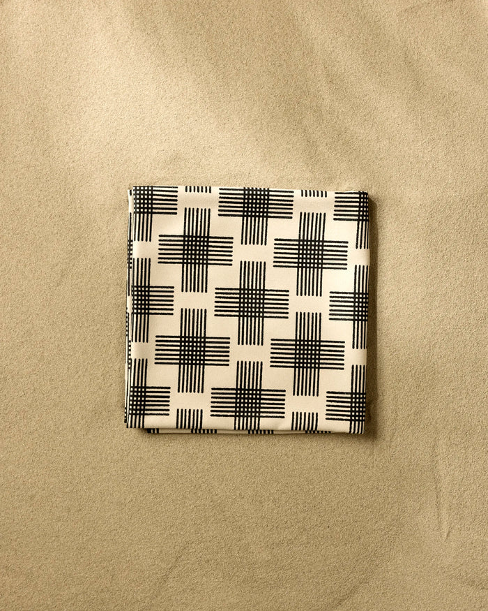 A folded Rosa Kichline Cross Hatch Micro-loop Beach Blanket in beige with a black geometric pattern rests on smooth sand.